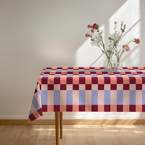 May include: A rectangular tablecloth featuring a geometric grid pattern in shades of deep burgundy, soft peach, and light periwinkle blue. The fabric drapes over a wooden table, which holds two clear glass vases containing small pink and white flowers.