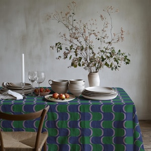 Può includere: Un tavolo da pranzo apparecchiato. Il tavolo è coperto da una tovaglia a fantasia blu, verde e viola. Ci sono piatti, ciotole, bicchieri e un vaso con fiori. Una sedia di legno è in primo piano.