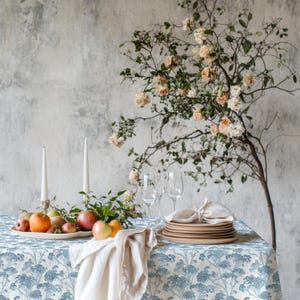 May include: A dining table set for a special occasion. The table is covered with a blue and white floral tablecloth and adorned with fruit, candles, plates, and wine glasses. A floral arrangement with peach and white flowers adds a natural touch.