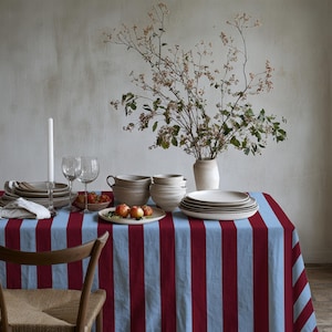 May include: A rectangular tablecloth featuring vertical stripes in alternating shades of light blue and deep burgundy red. The fabric is draped over a table, providing a base for a collection of ceramic bowls, plates, wine glasses, and a vase.