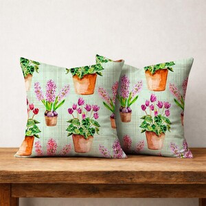 Op de afbeelding: Twee decoratieve kussens met een aquarel bloemenpatroon. Het ontwerp toont potplanten met roze en paarse bloemen en groen blad op een lichtgroene achtergrond. De kussens liggen op een houten oppervlak.