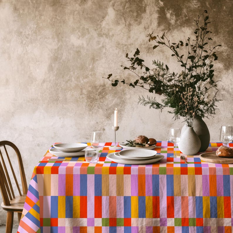 May include: A rectangular tablecloth featuring a vibrant, multi-colored geometric grid pattern with alternating vertical and horizontal stripes in shades of orange, red, blue, pink, yellow, and green, draped over a dining table with white plates.