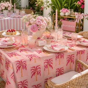 May include: A table setting featuring a pink tablecloth with a repeating palm tree pattern, topped with a floral centerpiece, pink taper candle, glassware, plates with pink napkins, a cake stand with cupcakes, and a sign labeled Aloha.