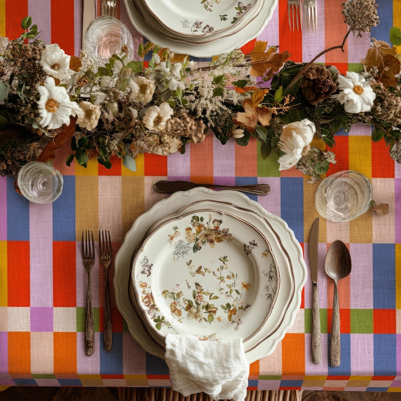 May include: A table setting featuring a white ceramic plate with a floral pattern, a white napkin, two forks, a knife, and a spoon. The setting is placed on a colorful plaid tablecloth with orange, purple, blue, and yellow blocks.