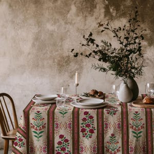 May include: A dining table set with a floral patterned tablecloth in cream, red, and green. The table is set with white plates, glasses, and a vase of flowers. A lit candle and wooden chair are also visible.