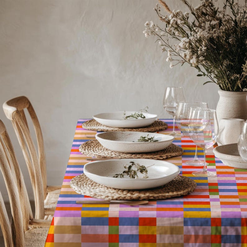 May include: A rectangular tablecloth featuring a vibrant, multi-colored grid pattern of orange, blue, pink, yellow, and green squares. The table is set with three white ceramic bowls on woven placemats, clear glassware, and a vase of dried flowers.