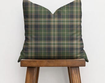 Puede incluir: Un cojín decorativo con un estampado de cuadros en tonos verdes, azul marino y beige. El cojín está colocado sobre un taburete de madera, mostrando su diseño. El diseño de cuadros añade un toque de estilo clásico a la decoración del hogar.