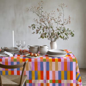 May include: A rectangular tablecloth featuring a vibrant, multi-colored geometric block pattern in shades of orange, pink, blue, yellow, and green. The table is set with white ceramic bowls, plates, wine glasses, a candle, and a vase with branches.