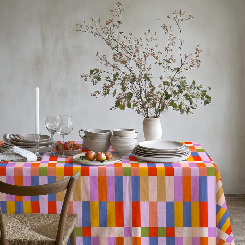 May include: A rectangular tablecloth featuring a vibrant, multi-colored geometric block pattern in shades of orange, pink, blue, yellow, and green. The table is set with white ceramic bowls, plates, wine glasses, a candle, and a vase with branches.