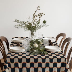 Può includere: Un tavolo da pranzo apparecchiato con una tovaglia fantasia blu navy, beige e arancione. Piatti bianchi, bicchieri trasparenti e un centrotavola floreale con verde e fiori bianchi sono disposti sul tavolo. Sedie in legno circondano il tavolo.
