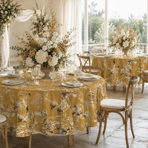 May include: A round table covered in a mustard yellow tablecloth featuring a white and dark brown toile de jouy pattern of pagodas and trees. The table is set with white plates, clear glassware, and a large floral centerpiece of white and gold flowers.