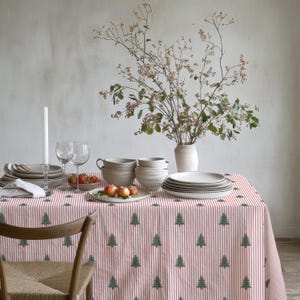 May include: A rectangular tablecloth featuring a pattern of vertical red stripes and small green pine trees. The fabric covers a dining table set with stacks of white ceramic plates, bowls, wine glasses, a white candle, and a bowl of apples.