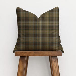 Puede incluir: Un coj&iacute;n cuadrado con un estampado de cuadros escoceses en tonos verde oliva, marr&oacute;n y beige. El coj&iacute;n est&aacute; colocado sobre un taburete de madera. El dise&ntilde;o de cuadros se forma con l&iacute;neas que se cruzan, creando un aspecto texturizado tradicional.