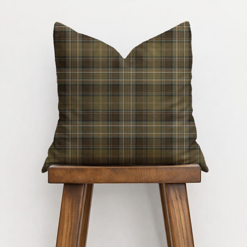 Puede incluir: Un coj&iacute;n cuadrado con un estampado de cuadros escoceses en tonos verde oliva, marr&oacute;n y beige. El coj&iacute;n est&aacute; colocado sobre un taburete de madera. El dise&ntilde;o de cuadros se forma con l&iacute;neas que se cruzan, creando un aspecto texturizado tradicional.