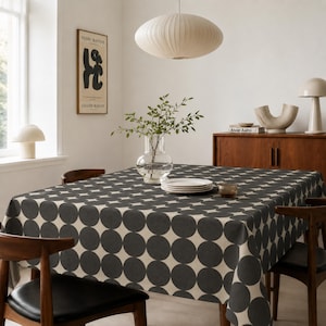 May include: A square tablecloth featuring a repeating pattern of large, dark charcoal grey circles on a light cream background, draped over a dining table. The fabric covers the top and sides of the table, showcasing a bold, geometric polka dot design.