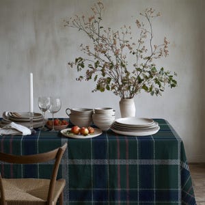Puede incluir: Un mantel de cuadros verde oscuro con finas rayas rojas y blancas, decorado con pilas de platos y cuencos de cerámica color crema, una vela blanca en un portavelas de cristal, dos copas de vino, un cuenco con bayas y un jarrón con ramas.