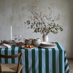 Puede incluir: Un mantel rectangular con rayas verticales en verde bosque oscuro y azul cielo claro. La tela cubre una mesa de comedor dispuesta con platos de cerámica, cuencos, copas de vino, una vela blanca y un jarrón con ramas secas.