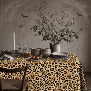 Könnte beinhalten: Eine rechteckige Tischdecke mit einem sich wiederholenden Muster aus schwarzen und hellbraunen Leopardenflecken, durchsetzt mit orangefarbenen menschlichen Schädeln. Der Stoff bedeckt einen Tisch mit Stapeln aus Keramikschalen, Tellern, Weingläsern und einer weißen Kerze.