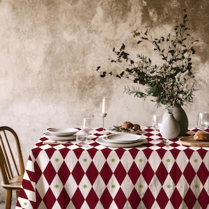 May include: A rectangular tablecloth featuring a repeating diamond pattern in deep red and white, accented with small green star-like motifs at the intersections. The fabric covers a dining table set with white ceramic plates, glassware, and a vase.