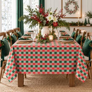 Puede incluir: Una mesa de comedor rectangular cubierta con un mantel de cuadros rojos y verdes. La mesa está puesta con platos blancos, vasos transparentes y servilletas verdes. Un gran jarrón blanco lleno de ramas de hoja perenne, bayas rojas y flores blancas se encuentra en el centro.