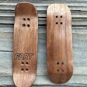 May include: Two wooden fingerboards with a natural wood grain finish. One board has the word "FART" etched into the surface. Each board has four sets of pre-drilled holes for mounting trucks. The boards are designed for finger skateboarding.