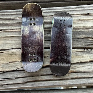 May include: Two finger skateboards with a dark brown and black design. One board has the word "FART" printed on it. Each board has four holes for the trucks.