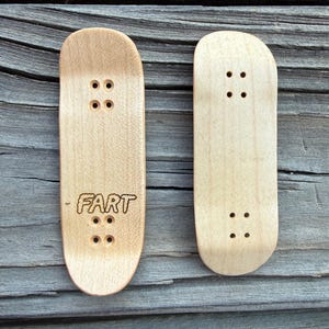 May include: Two miniature wooden finger skateboards. One board has the word "FART" etched into the wood. Both boards have four sets of pre-drilled holes for the trucks. The wood has a natural, light brown color.