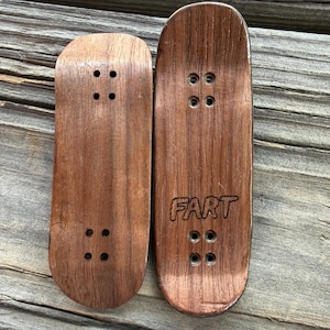 May include: Two small, wooden fingerboards with a dark brown finish. One board has the word "FART" etched into the wood. Each board has four sets of pre-drilled holes for mounting trucks. The boards are displayed on a wooden surface.