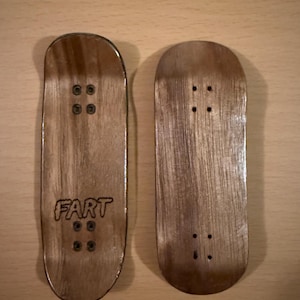 May include: Two wooden fingerboards with a natural wood grain finish. One board has the word "FART" etched into the wood. Each board has four small holes for the trucks and hardware. The boards are designed for finger skateboarding.