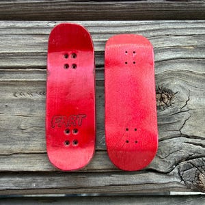 May include: Two red finger skateboards with black hardware. One board has the word "FART" in black letters. The boards are displayed on a weathered wooden surface.