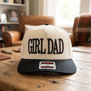 May include: A beige and black baseball cap with the words "GIRL DAD" embroidered in bold black letters. The cap has a flat brim and a small tag on the front.