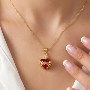 May include: A gold-colored necklace with a heart-shaped pendant. The pendant is red with gold accents and small clear stones. The necklace is worn over a white collared shirt.