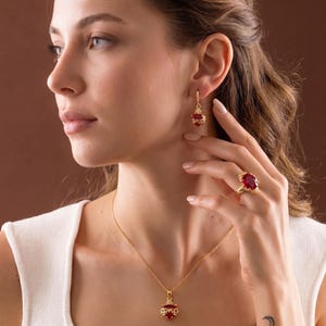 Op de afbeelding: Een goudkleurige sieradenset met een ketting, oorbellen en een ring, allemaal versierd met levendige rode edelstenen. De ring zit aan een vinger en de oorbellen hangen aan het oor. De hanger van de ketting en de oorbellen hebben een vergelijkbaar ontwerp.