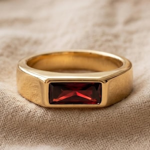 May include: A gold-toned ring featuring a single rectangular, deep red gemstone set horizontally in the center. The band is smooth and polished, with a flat profile that highlights the vibrant, faceted red stone against the metallic gold finish.