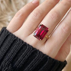Puede incluir: Un anillo de oro con una gran piedra preciosa de color rojo intenso, de corte esmeralda rectangular y sujeta por cuatro garras. El anillo se lleva en un dedo y la banda es de oro amarillo pulido, con un diseño sencillo y limpio.