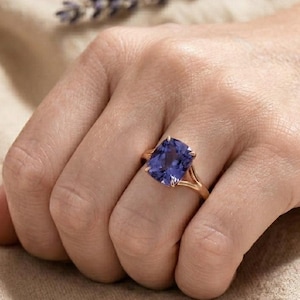 May include: A gold ring featuring a large, cushion-cut purple gemstone set in a four-prong mounting. The band is thin and polished, showcasing a vibrant violet stone that sits prominently on the finger, reflecting light across its faceted surface.