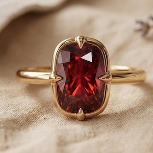 Può includere: Un anello in oro con una grande pietra preziosa rosso intenso dal taglio a cuscino rettangolare, incastonata in una montatura a quattro griffe. La fascia è liscia e lucida, con un design essenziale che esalta il colore vivido e la superficie sfaccettata.