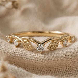 Pode incluir: Um anel de ouro com uma banda em forma de V adornada com pequenas pedras redondas transparentes e motivos de folhas gravados. O design incorpora folhas de metal e pedras brilhantes alternadas, criando um aspeto delicado e simétrico.