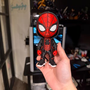 Puede incluir: Una figura de Spider-Man impresa en 3D. La figura es predominantemente roja y negra, con detalles de ojos blancos. Lleva una chaqueta negra y zapatillas deportivas. La figura se sostiene en una mano.