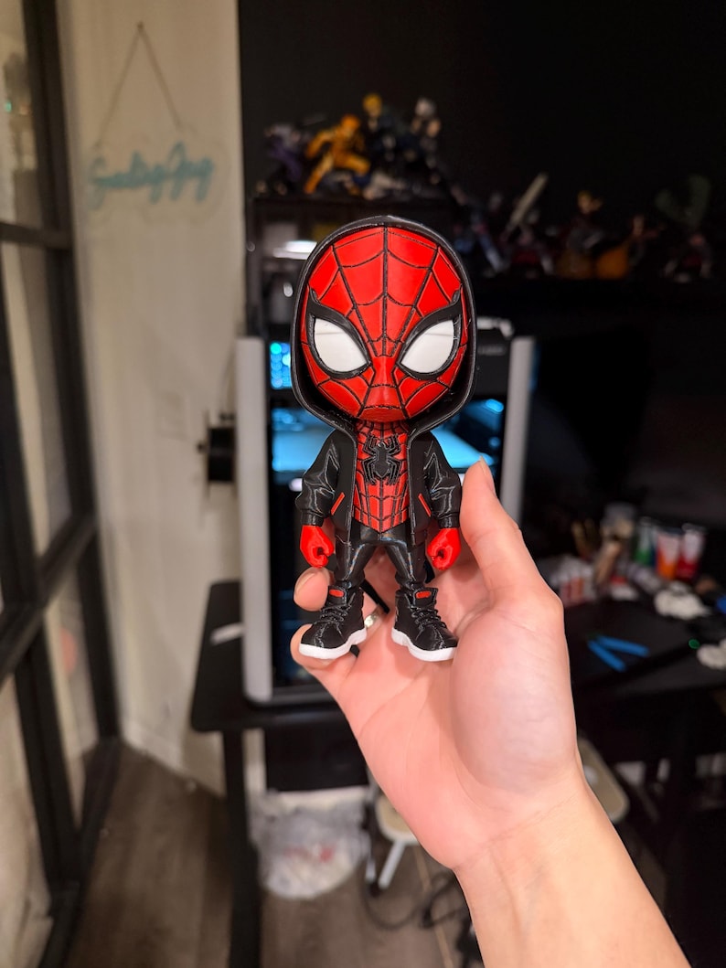 Puede incluir: Una figura de Spider-Man impresa en 3D. La figura es predominantemente roja y negra, con detalles de ojos blancos. Lleva una chaqueta negra y zapatillas deportivas. La figura se sostiene en una mano.