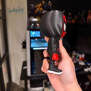Puede incluir: Una figura impresa en 3D de un superh&eacute;roe con un traje negro y detalles en rojo. La figura tiene una capucha negra, una m&aacute;scara roja y negra y zapatillas negras con suelas blancas. La figura se sostiene en una mano.
