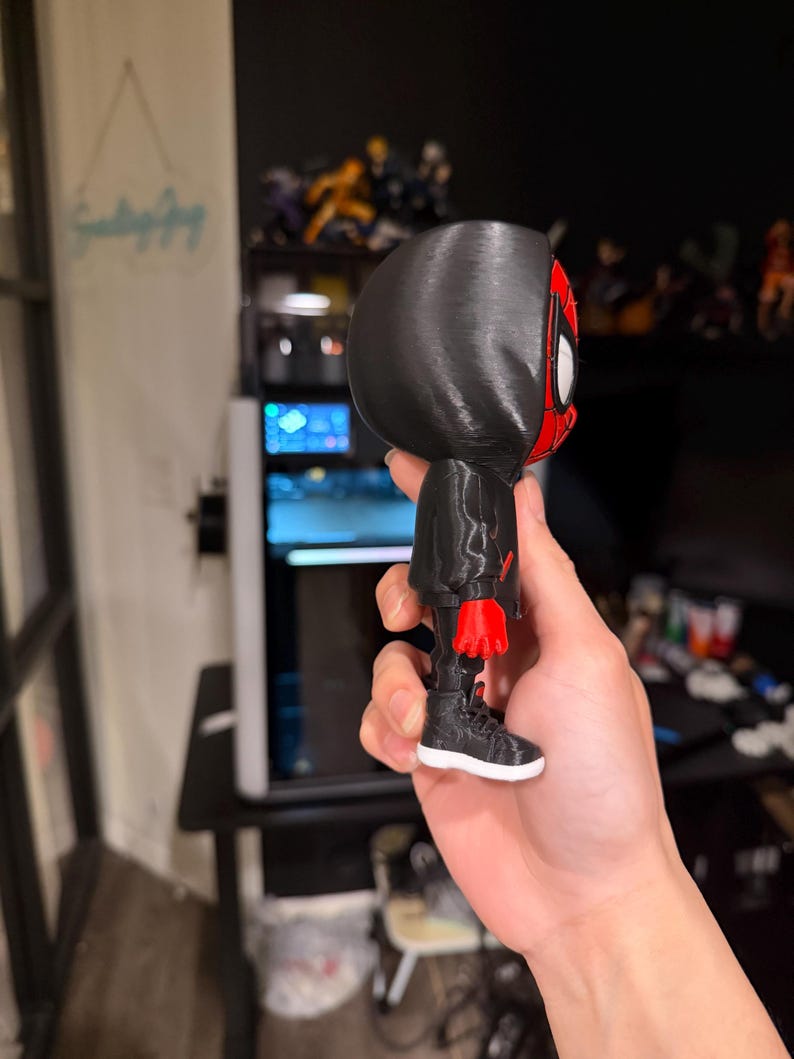 Puede incluir: Una figura impresa en 3D de un superh&eacute;roe con un traje negro y detalles en rojo. La figura tiene una capucha negra, una m&aacute;scara roja y negra y zapatillas negras con suelas blancas. La figura se sostiene en una mano.