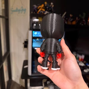 Puede incluir: Una figura negra de un boxeador con chaqueta negra, pantalones negros, guantes de boxeo rojos y botas negras con suelas blancas. La figura se sostiene en una mano, con un fondo borroso de una computadora y otras figuras.