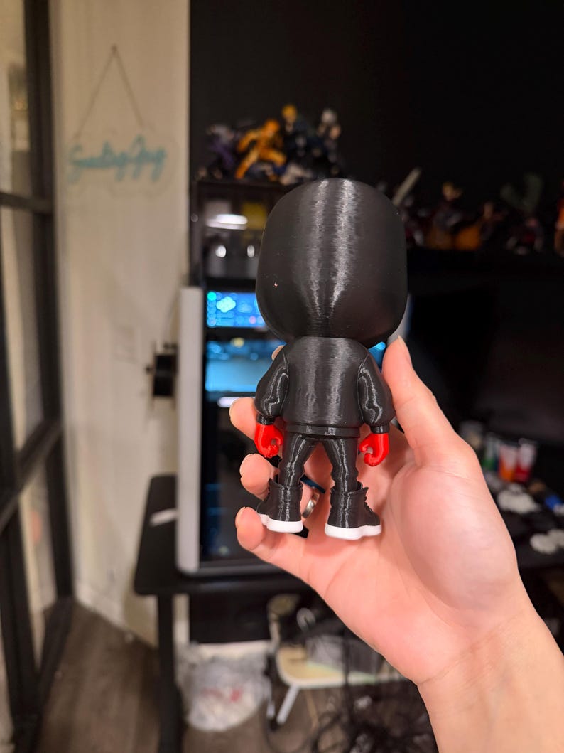 Puede incluir: Una figura negra de un boxeador con chaqueta negra, pantalones negros, guantes de boxeo rojos y botas negras con suelas blancas. La figura se sostiene en una mano, con un fondo borroso de una computadora y otras figuras.