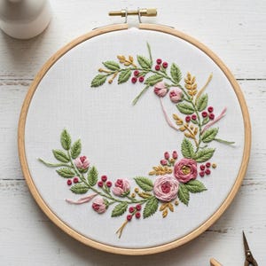 Puede incluir: Un bastidor de bordado floral con un diseño de semicírculo de rosas rosas, hojas verdes y bayas rojas. El bordado está colocado en un bastidor de madera. Una bobina de hilo rosa y unas tijeras pequeñas están cerca.