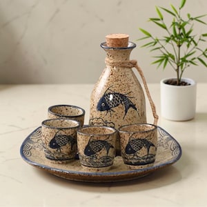 May include: A ceramic sake set featuring a bottle with a cork stopper and a rope handle, a serving tray, and four small cups. The set is beige with blue fish illustrations and wave patterns.