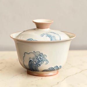 May include: A ceramic tea bowl with a lid, decorated with a wave pattern in blue and grey. The bowl has a light beige body with a pinkish-brown rim and base. The lid features a wave design and a pinkish-brown handle.