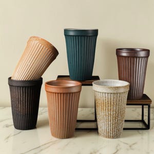 Puede incluir: Colección de seis tazas de café de cerámica en varios colores, incluyendo negro, marrón, beige y verde azulado. Cada taza tiene una textura acanalada y una forma cilíndrica. Algunas tazas se exhiben en pequeñas estanterías de madera.