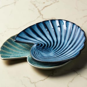 Op de afbeelding: Een keramieken schaal in de vorm van een nautilusschelp met een glanzende afwerking. Het bovenste gedeelte heeft diepblauwe tinten met stralende ribbels, terwijl het onderste gedeelte overgaat in een lichtere blauwgroene kleur met textuur.