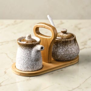 May include: A ceramic condiment set with a speckled white and brown glaze. The set includes a small pitcher with a spout, a lidded jar, and a spoon, all on a bamboo tray with a handle. The set is designed for serving sauces or spices.